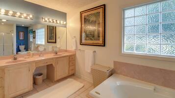A bathroom with a double vanity, a large mirror with lights, a bathtub, and a window with frosted glass.