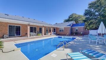 A rectangular swimming pool with a clear blue water surface, surrounded by a brick building with large windows and a covered patio area with lounge chairs and a table.