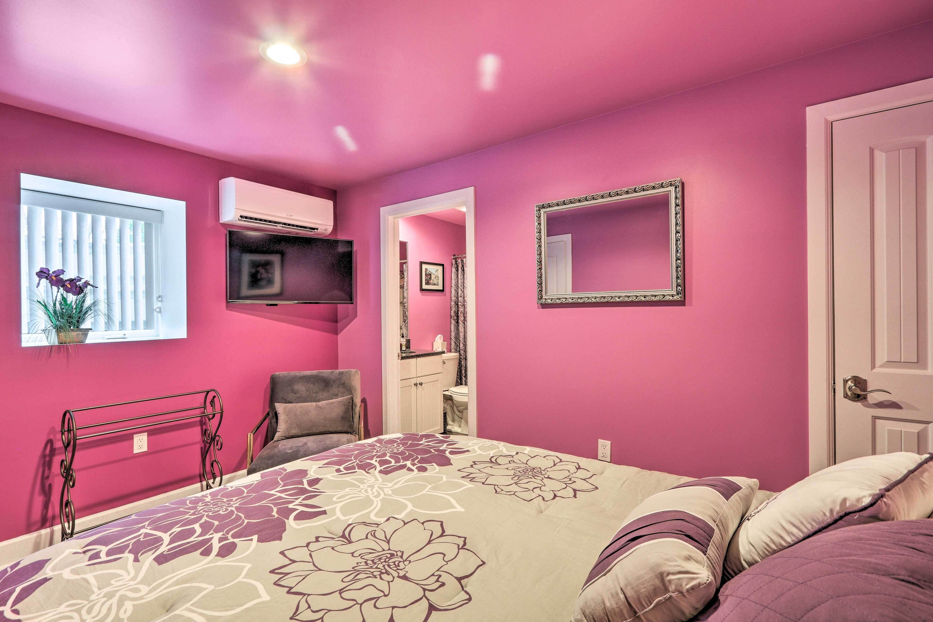 Un dormitorio con pared rosa, una cama con ropa de cama floral, una silla, un televisor y un equipo de aire acondicionado.