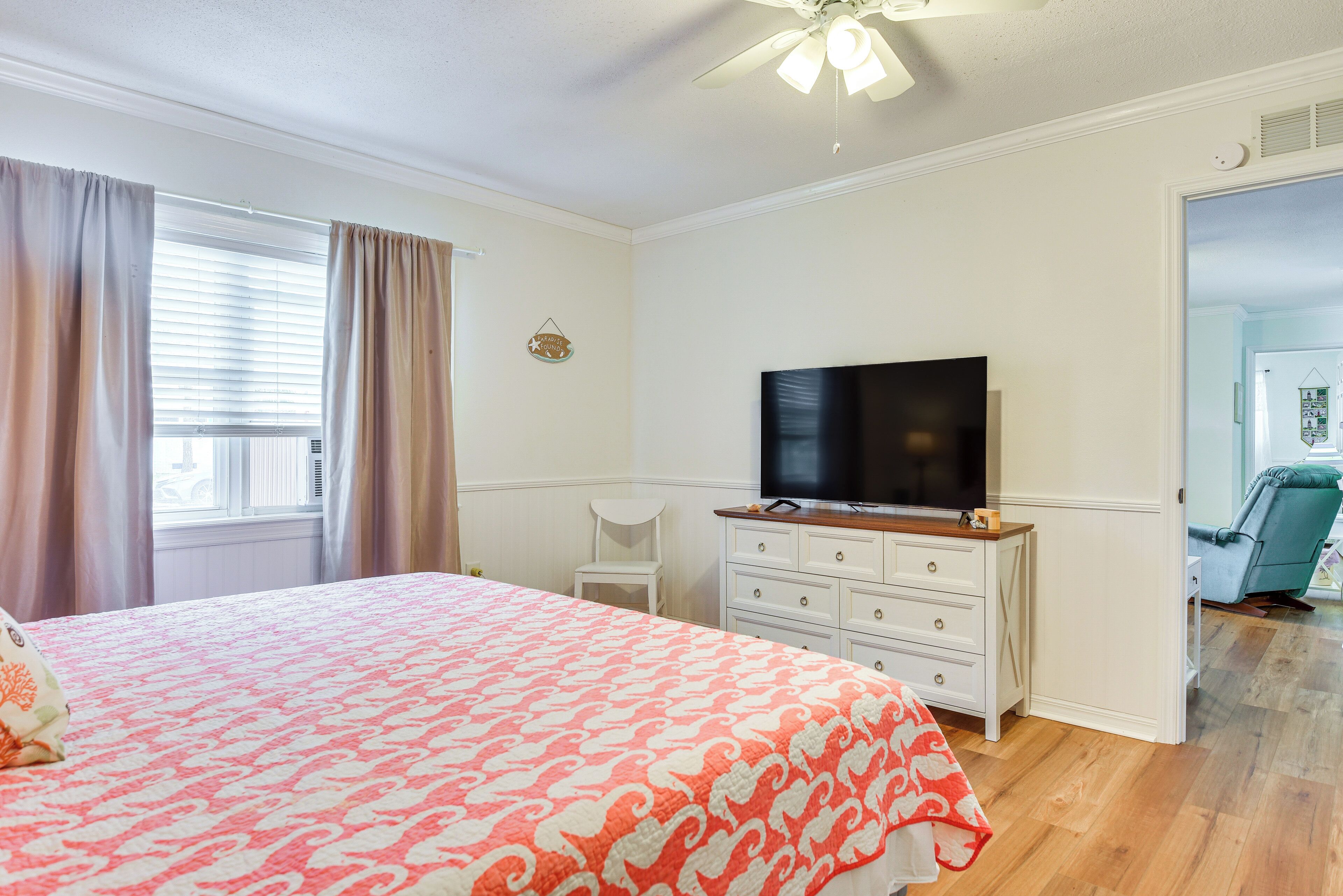 Quarto com cama, televisão sobre um penteadeira, uma cadeira e janela com cortinas.