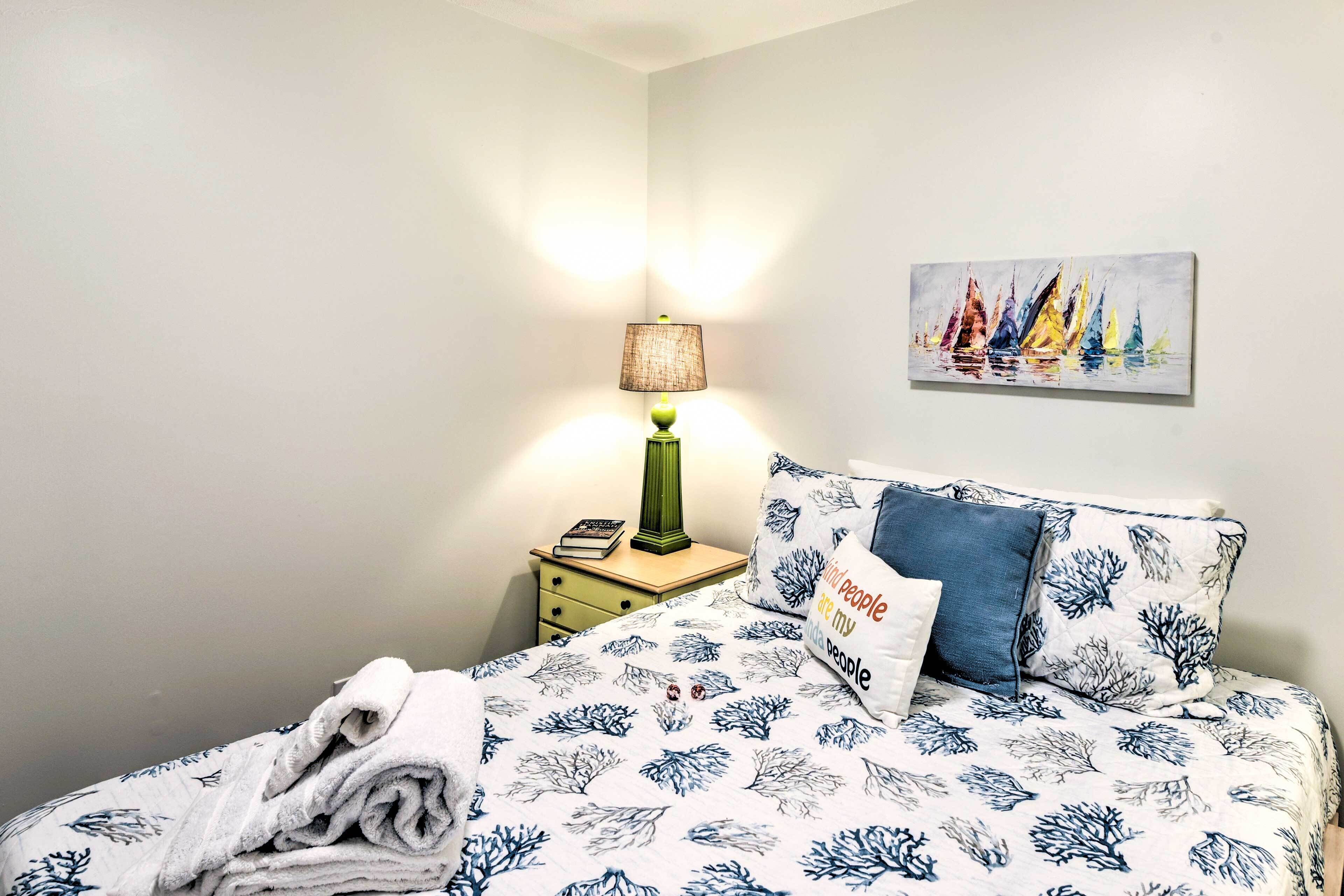 A neatly made bed with a floral-patterned bedspread, a folded towel on the side, a green lamp on a bedside table, and a painting of sailboats on the wall.