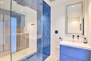 A modern bathroom with a glass shower enclosure, a blue-tiled shower wall, a white sink with a mirror, and a blue cabinet.