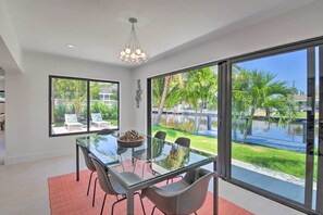 Uma sala de jantar moderna com mesa e cadeiras de vidro, janelas amplas com vista para uma piscina e vegetação.