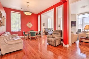 A living room with a red accent wall, a wooden floor, a beige sofa, a brown armchair, and a dining area with a table and chairs.