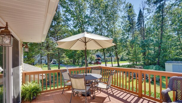 Um deck de madeira com mesa e cadeiras, um guarda-sol e vista para uma área arborizada.