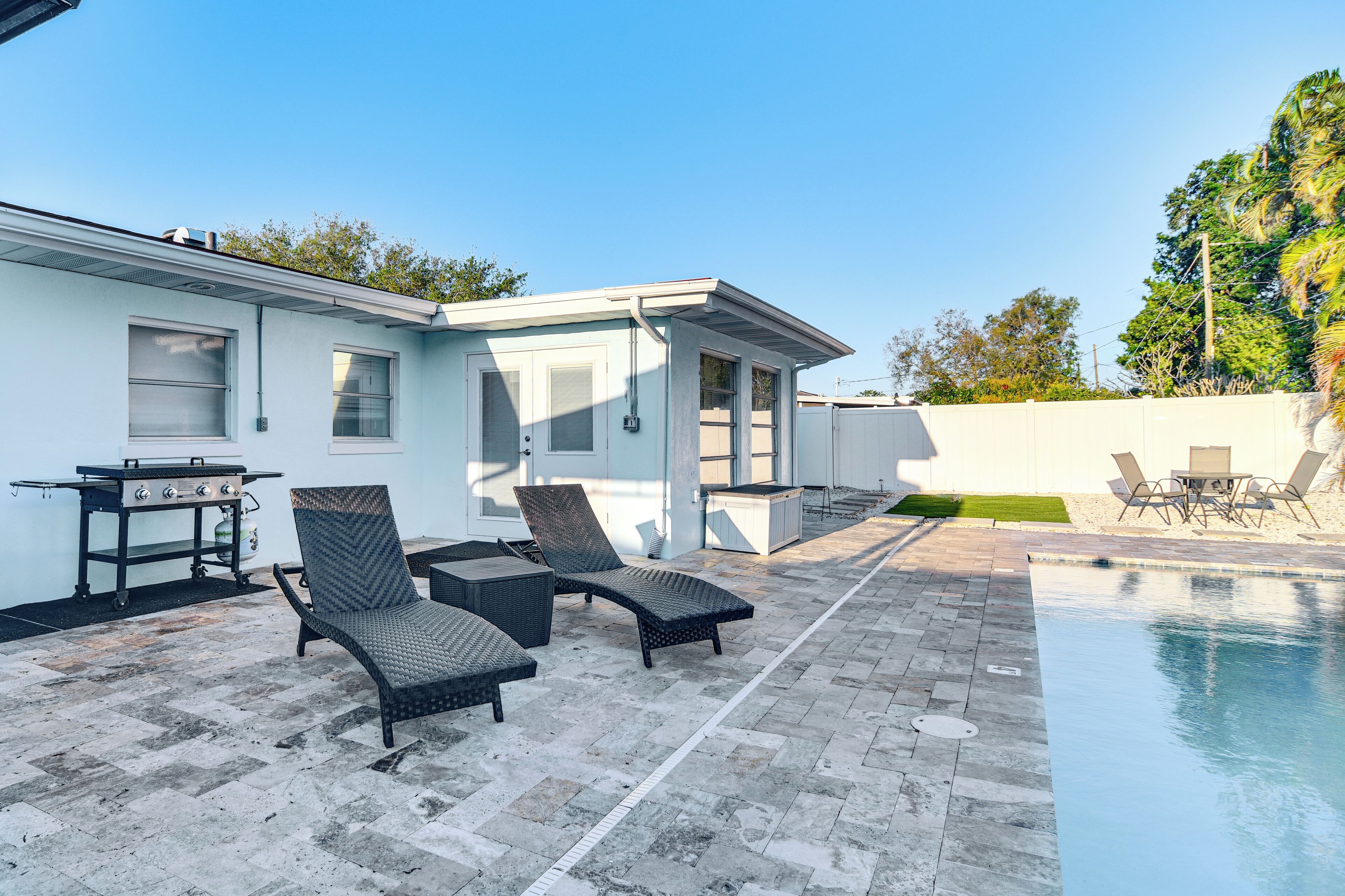 Een modern terras buiten met een zwembad, lounge stoelen, een barbecue en een kleine tafel.
