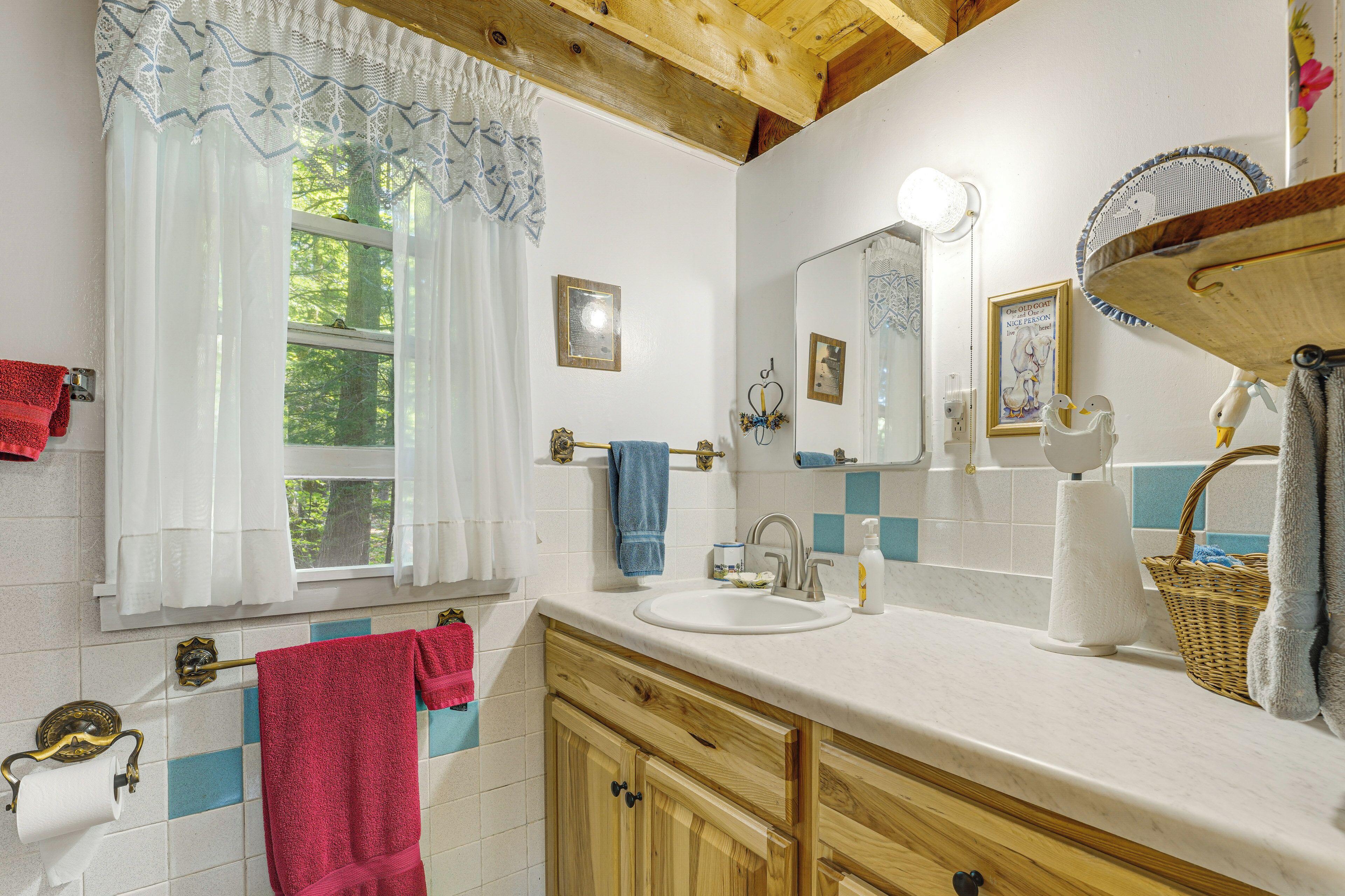 A bathroom with a wooden ceiling, a window with white curtains, a sink with a mirror, and a towel rack.