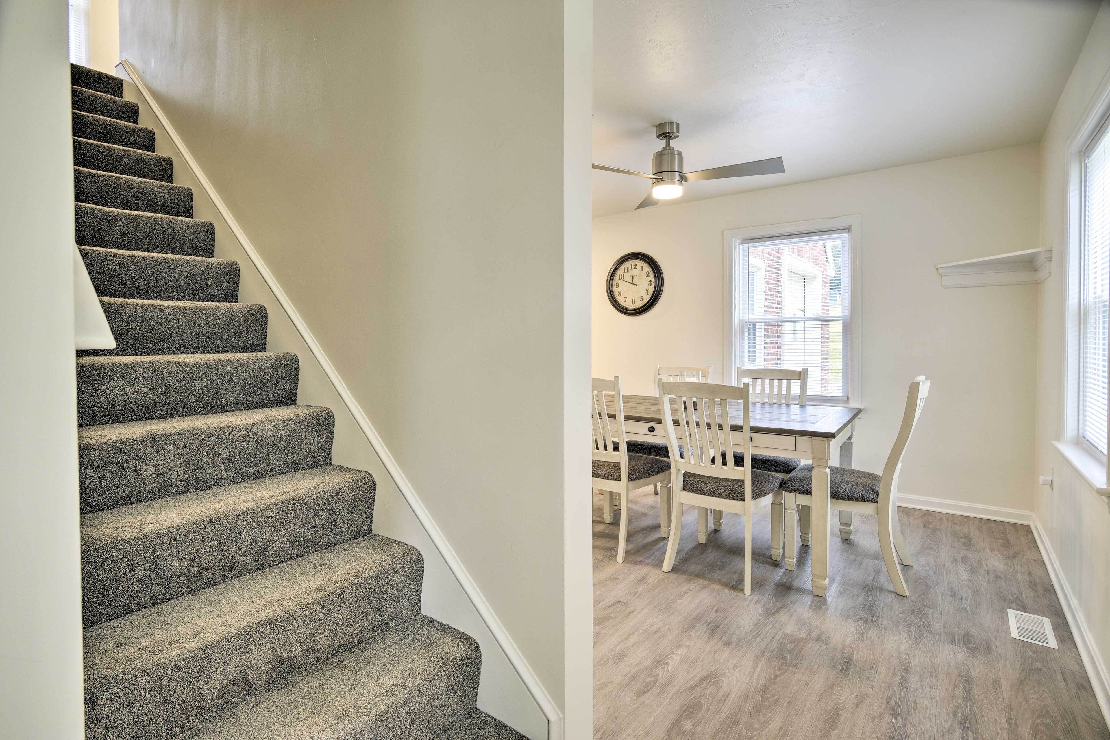 A staircase with carpeted steps, a dining area with a table and chairs, a clock on the wall, and a ceiling fan.