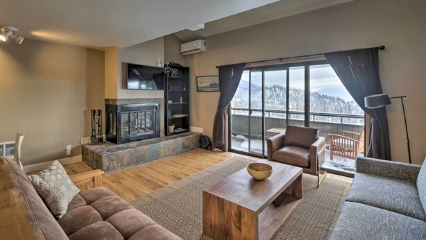A modern living room with a fireplace, comfortable seating, and a large window offering a view of the outdoors. - Whitefish Mountain Ski-in/out Condo w/ Views! (Whitefish)