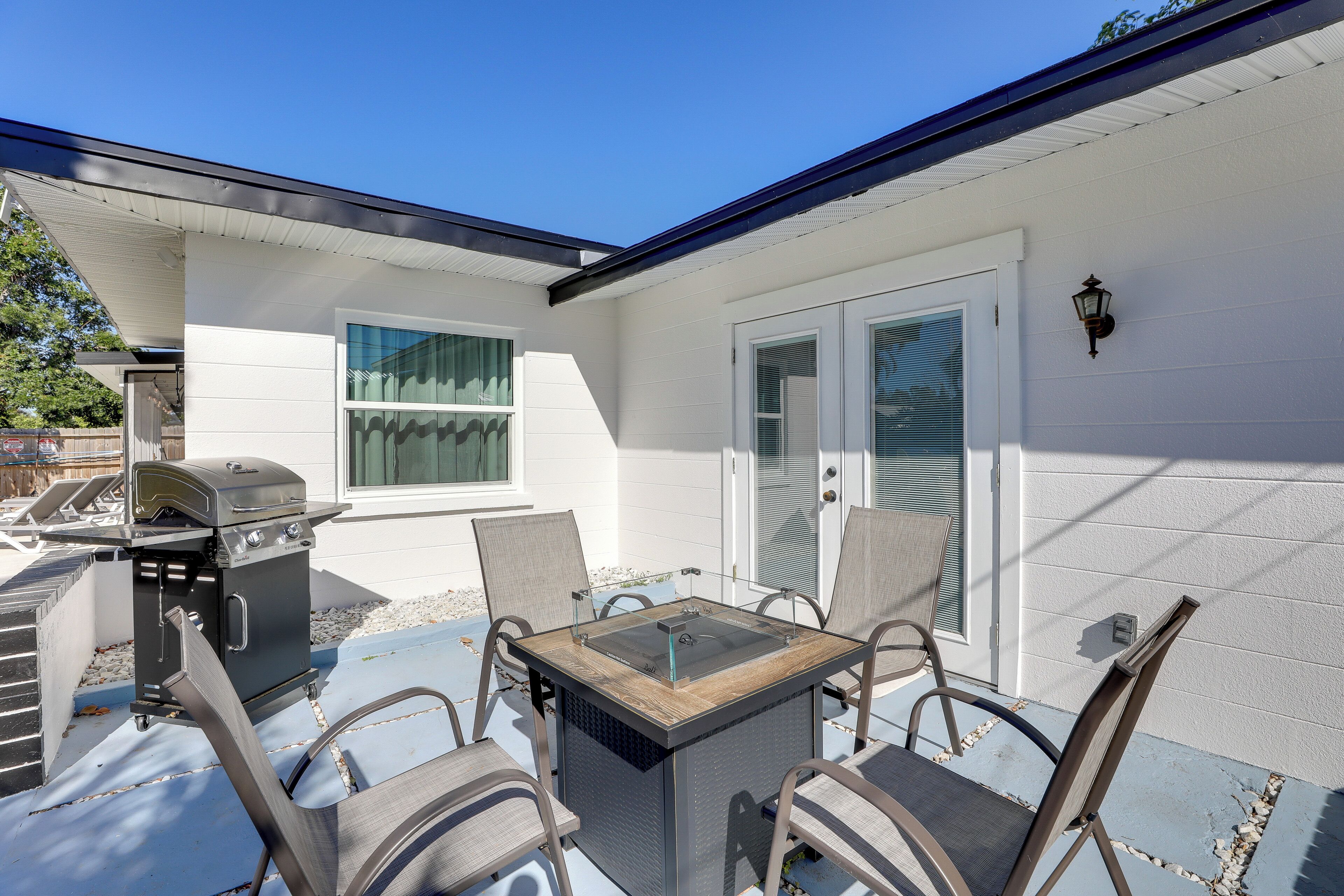 Een terras buiten met een barbecue, een tafel met glazen blad en vier stoelen.