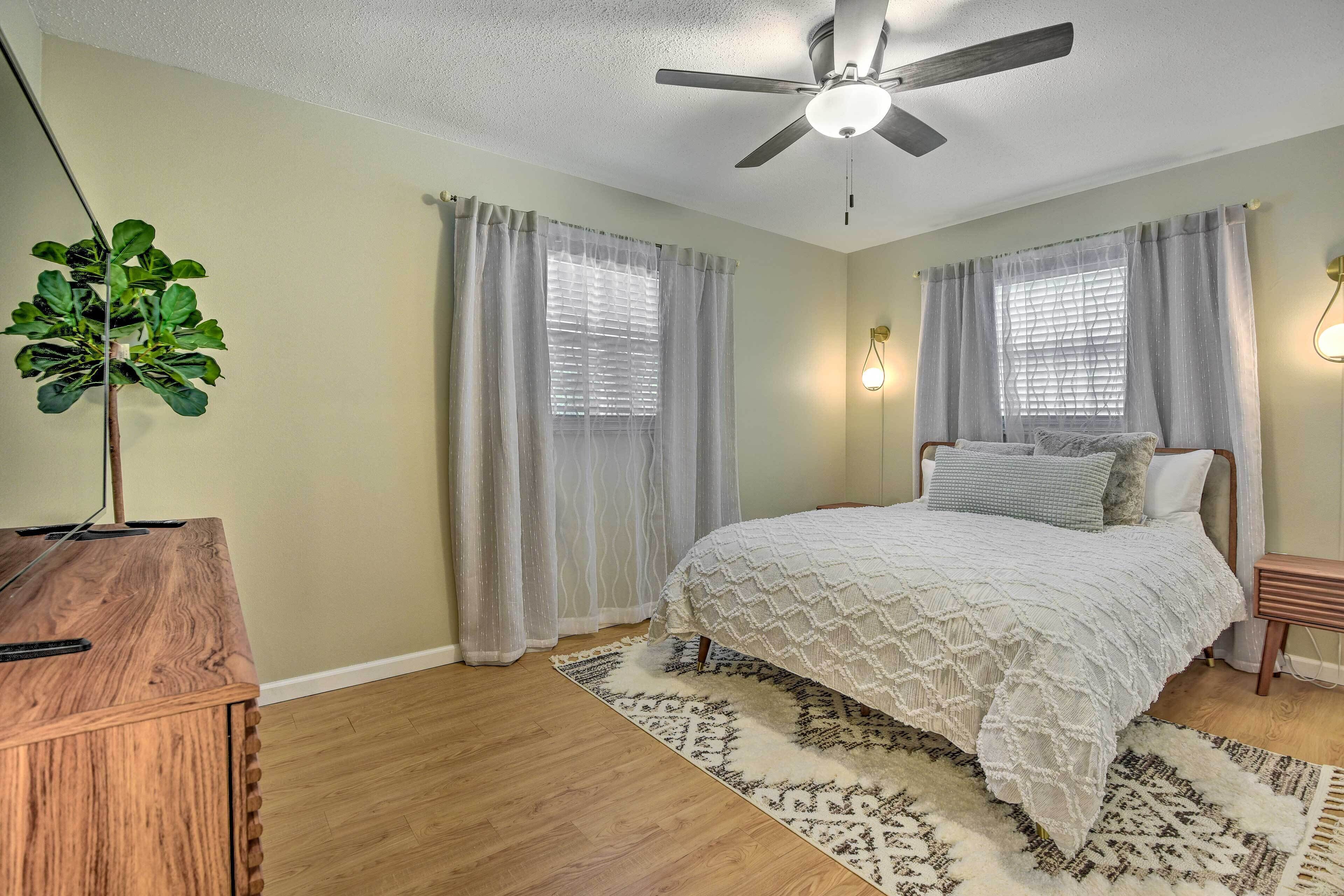 A bedroom with a bed, curtains, a ceiling fan, a plant, and a wooden desk.