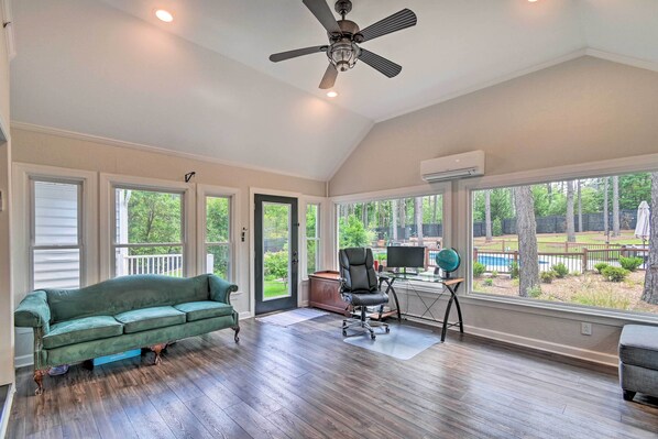 A spacious room with a green sofa, a black office chair, and a large window overlooking a backyard with a pool. - Peaceful Southern Pines Home w/ Pool + Yard! (Southern Pines)