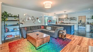 A modern living room with a sectional sofa, a wooden coffee table, and a colorful rug. There is a bookshelf with books and decorative items, and a kitchen area with white cabinets and a blue island. - Peaceful Southern Pines Home w/ Pool + Yard! (Southern Pines)