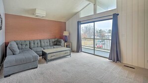 A living room with a sectional sofa, a small table, and a lamp. There is a large window with curtains and a view of the outside.
