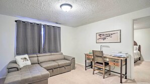 A room with a gray sectional sofa, a wooden desk with a chair, a framed picture on the wall, and a bed in the background.