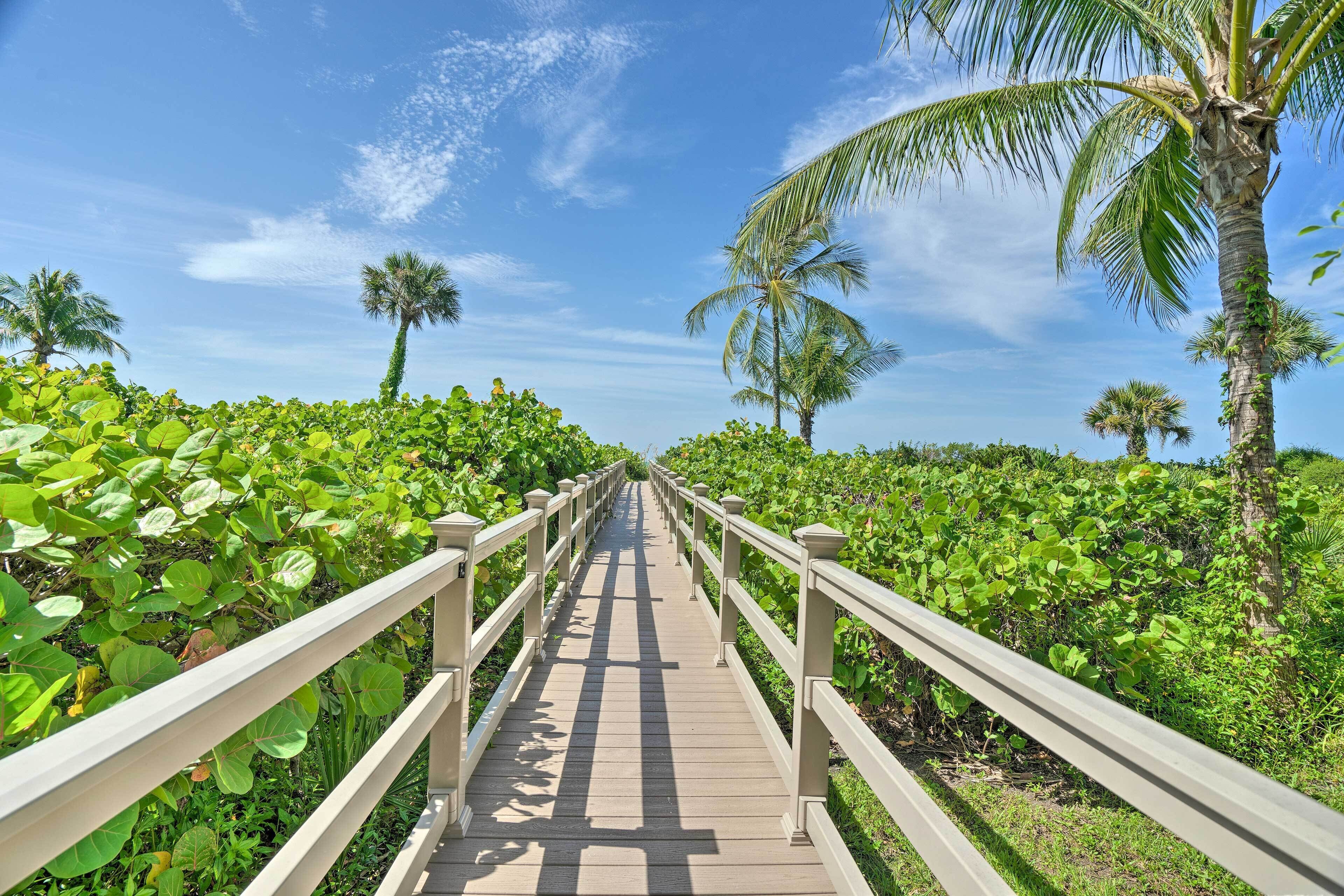 Un paseo marítimo de madera con barandillas blancas, rodeado de abundante follaje y palmeras.