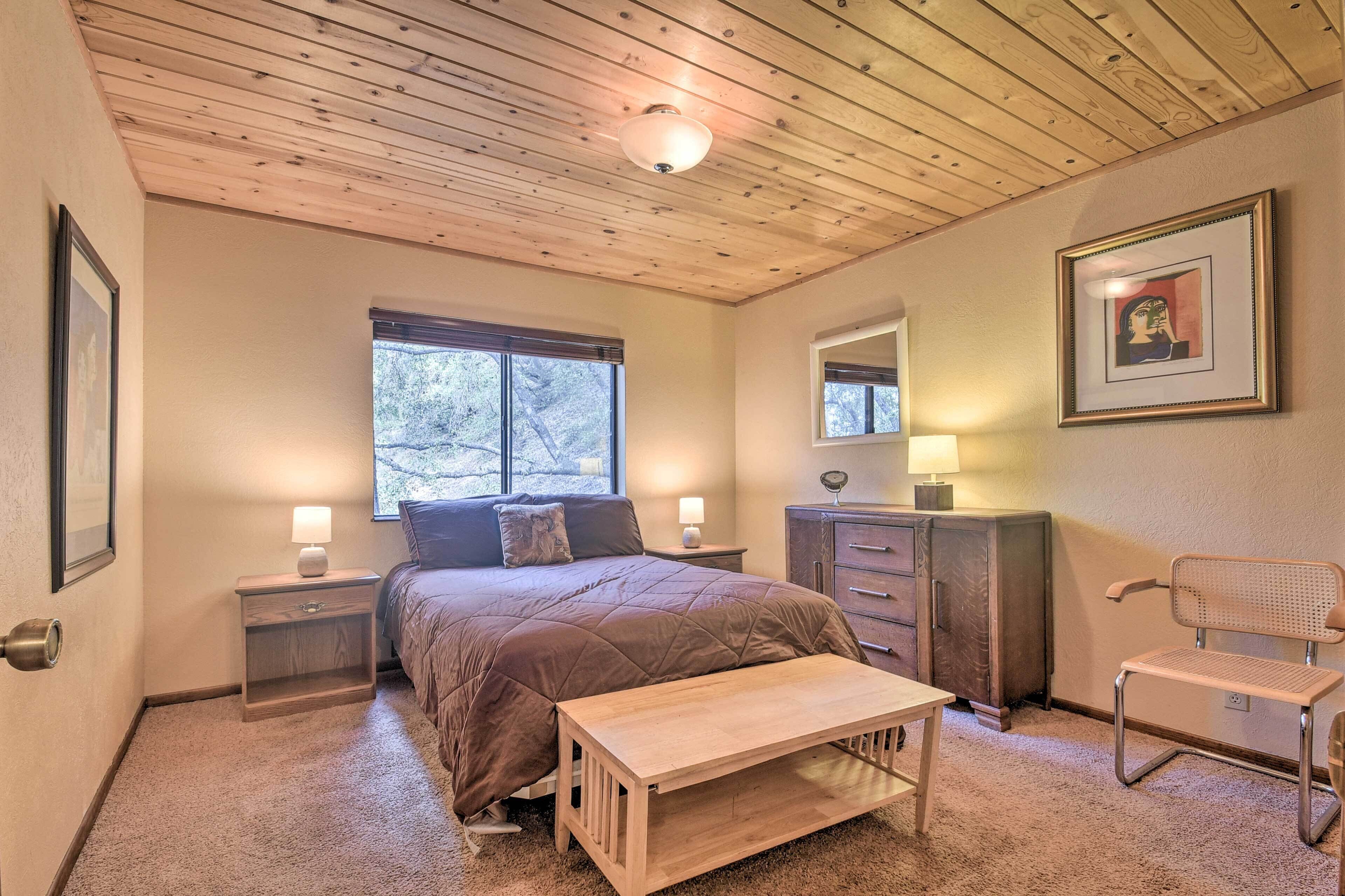 A bedroom with a wooden ceiling, a bed with a brown quilt, a nightstand with a lamp, a dresser, a chair, and a window with a view of trees.