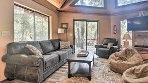 A cozy living room with a leather sofa, a coffee table, and a bean bag. Large windows offer views of trees. - Updated 'tree House' Pine Mtn Club Cabin by Trails (Pine Mountain Club)