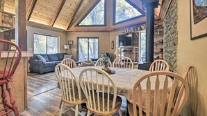 A dining area with a high ceiling, wooden beams, and large windows. There is a fireplace, a bookshelf, and a sofa in the background. - Updated 'tree House' Pine Mtn Club Cabin by Trails (Pine Mountain Club)