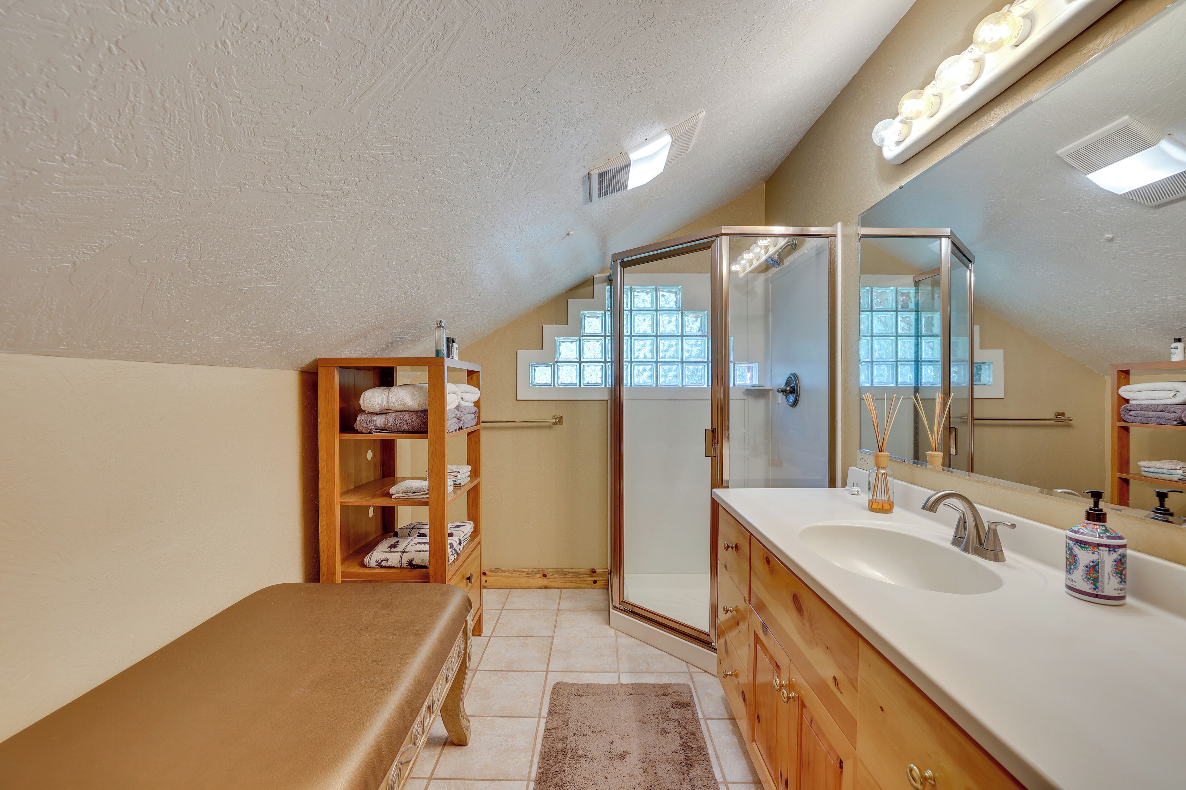A bathroom with a shower, a sink, and a mirror. There is a wooden shelf with towels and a rug on the floor.