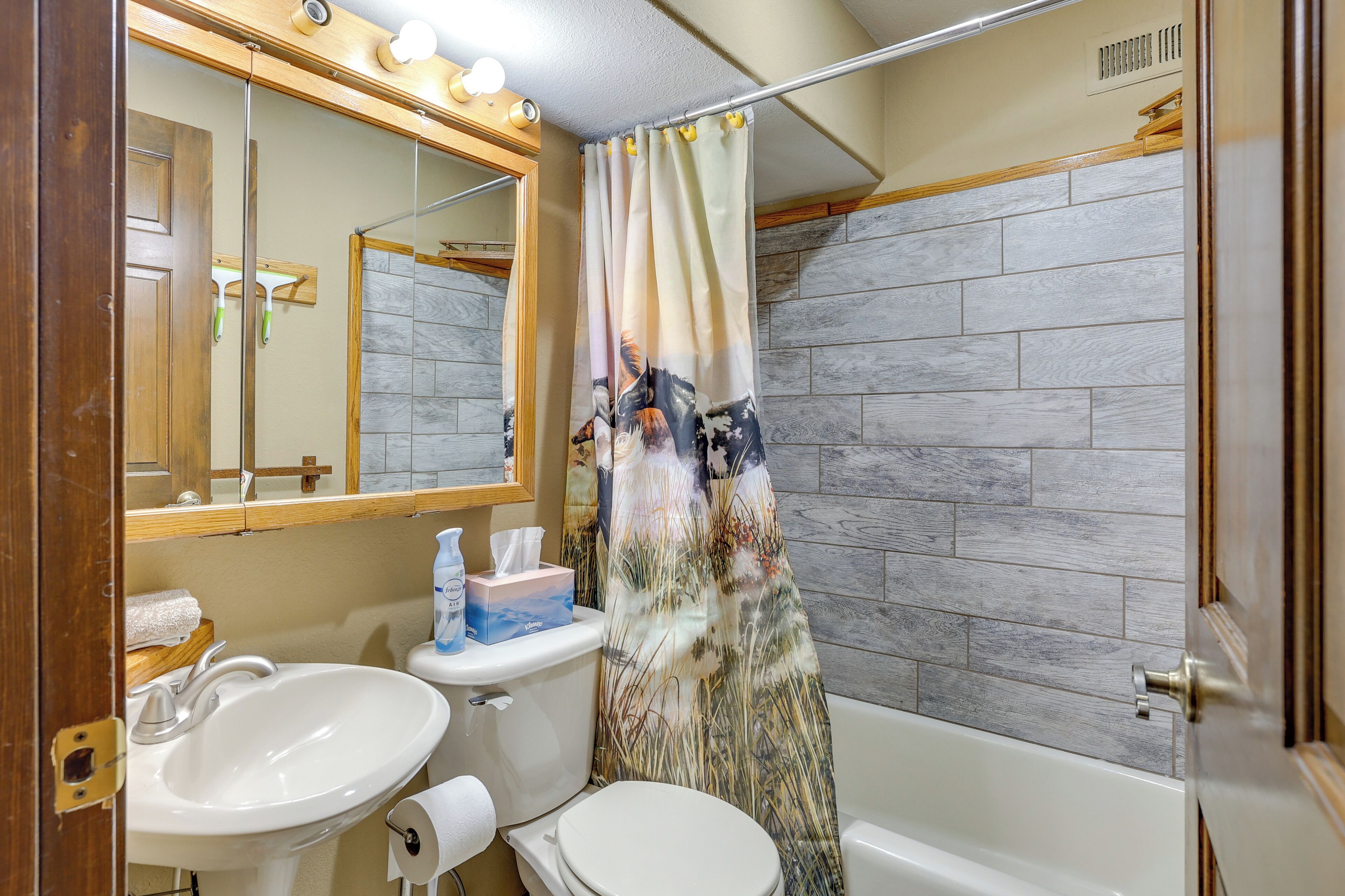 A bathroom with a white sink, toilet, and bathtub. There is a mirror above the sink and a shower curtain with a floral pattern. The walls are tiled with a gray stone-like material.
