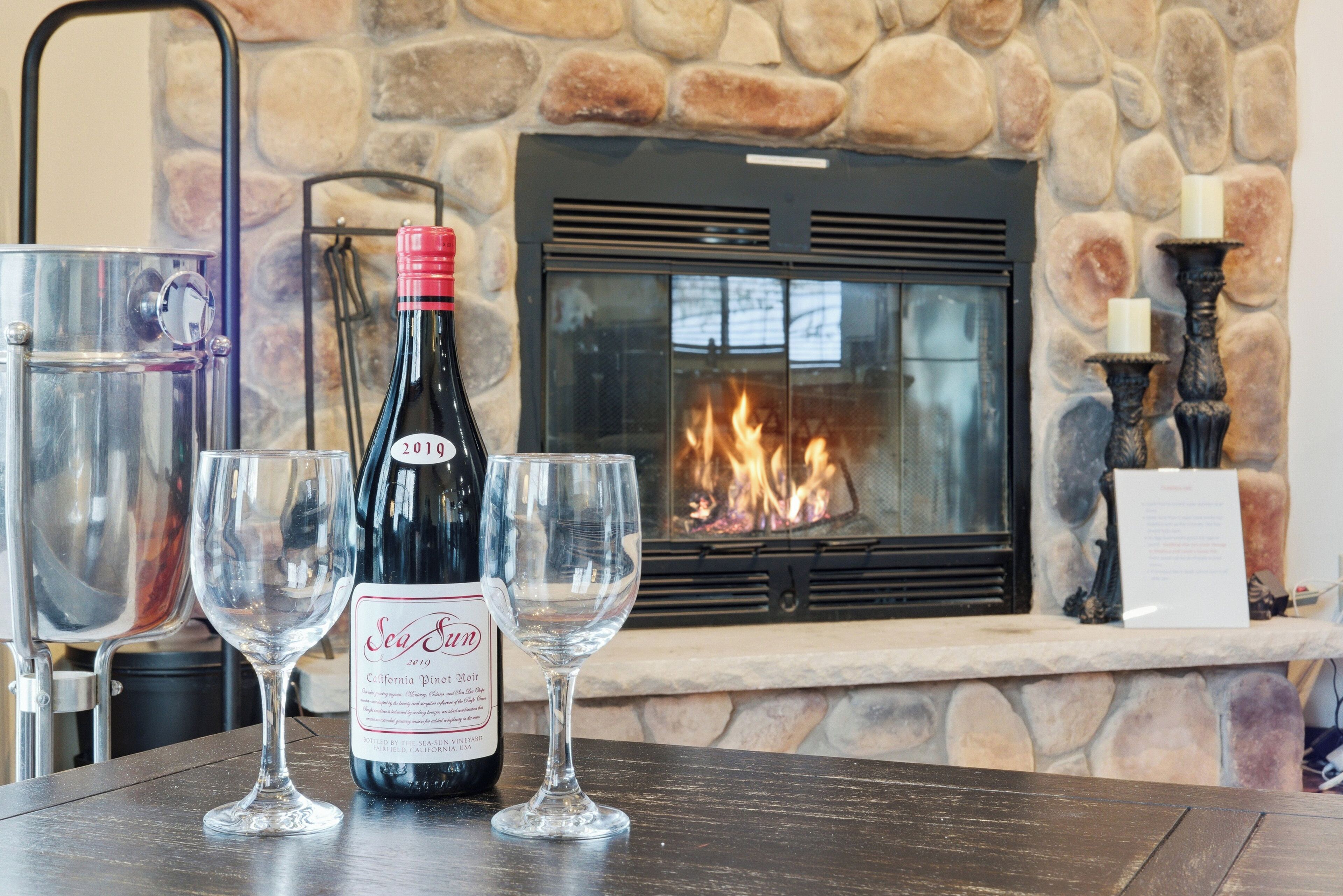 Une table avec une bouteille de vin et deux verres, une cheminée avec un mur en pierre et un présentoir métallique pour les verres.