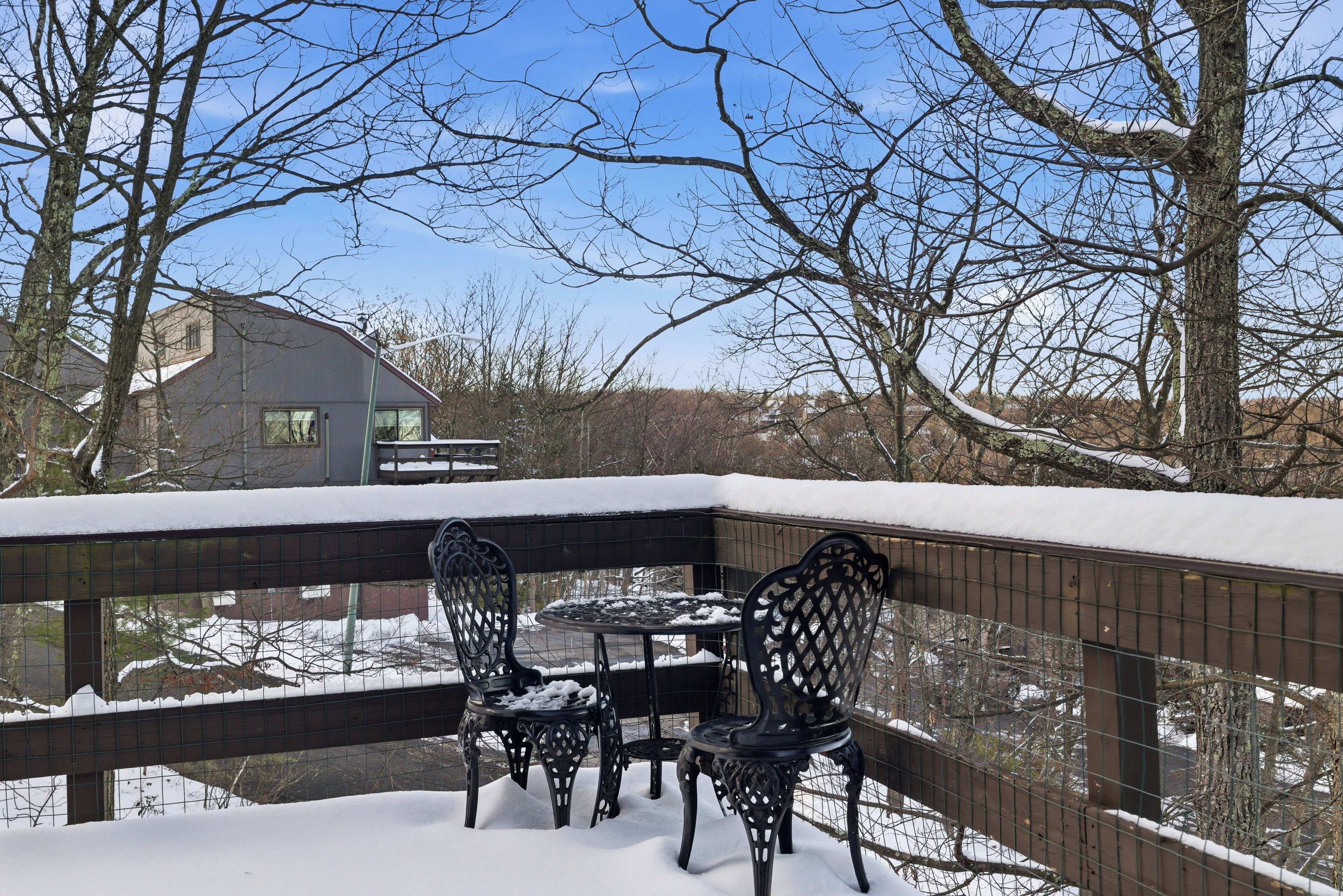 A snow-covered balcony with a table and chairs, a metal fence, and leafless trees in the background.