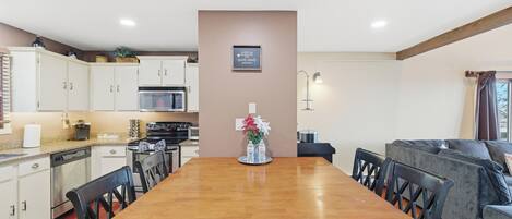 A modern kitchen with a wooden dining table, white cabinets, and a gray sofa in the living area.
