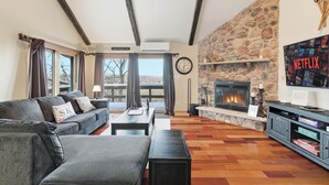 A cozy living room with a stone fireplace, a sofa, a coffee table, and a TV stand with books. - Cozy Camelback, Pocono Retreat w/ Mountain Views! (Tannersville)