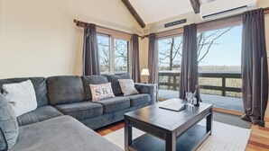 A modern living room with a grey sofa, a wooden coffee table, and large windows with curtains. - Cozy Camelback, Pocono Retreat w/ Mountain Views! (Tannersville)