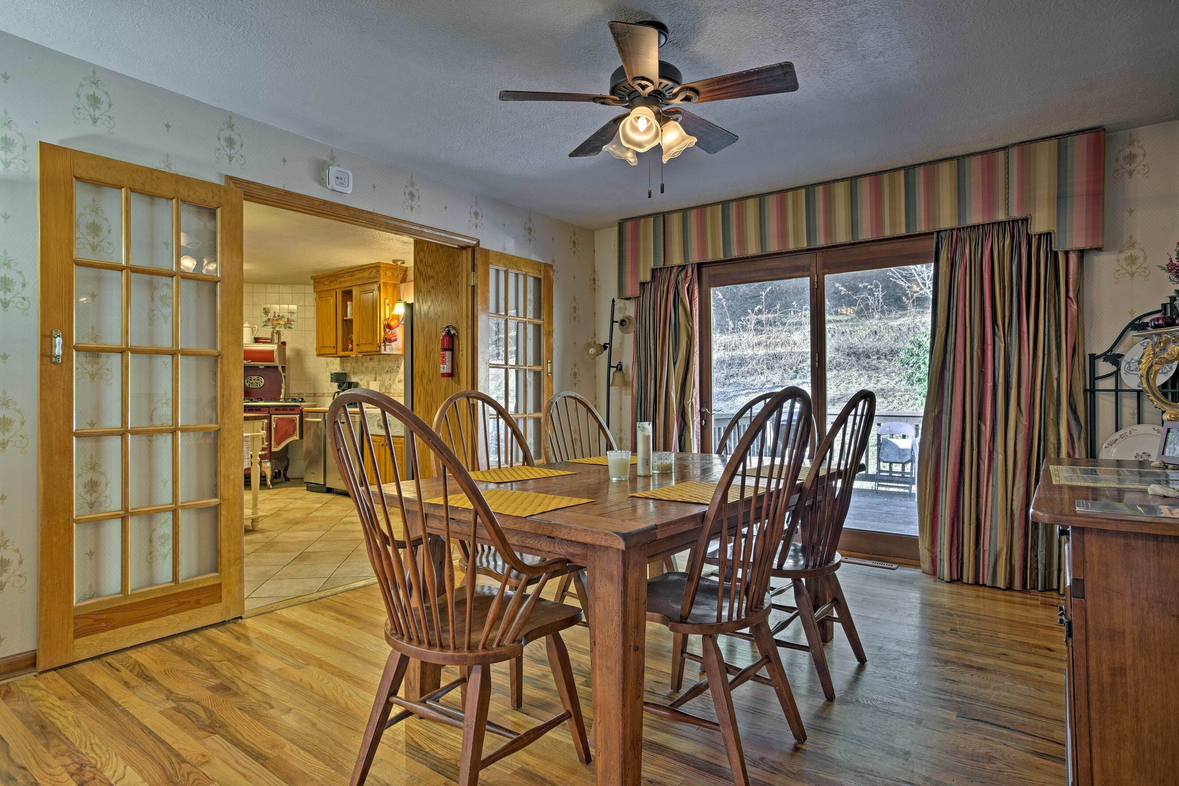 A dining area with wooden chairs and a table, a ceiling fan, and a view through a large window with curtains.