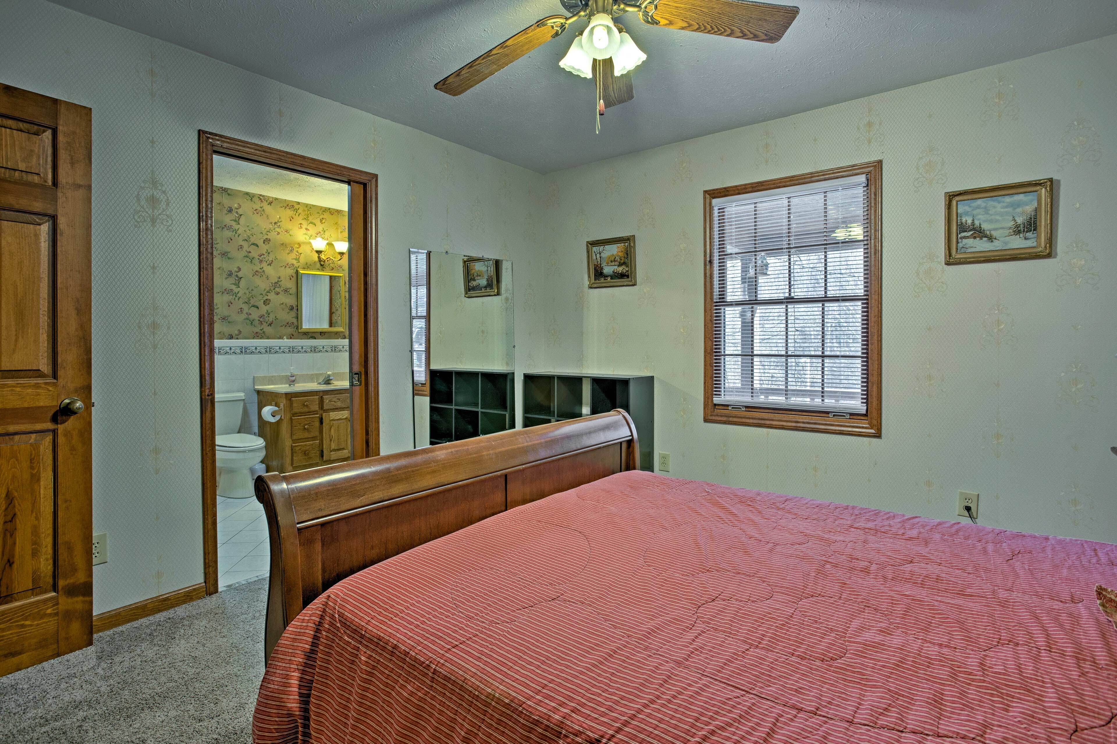 A bedroom with a wooden bed, a ceiling fan, a window with blinds, and a door leading to a bathroom.