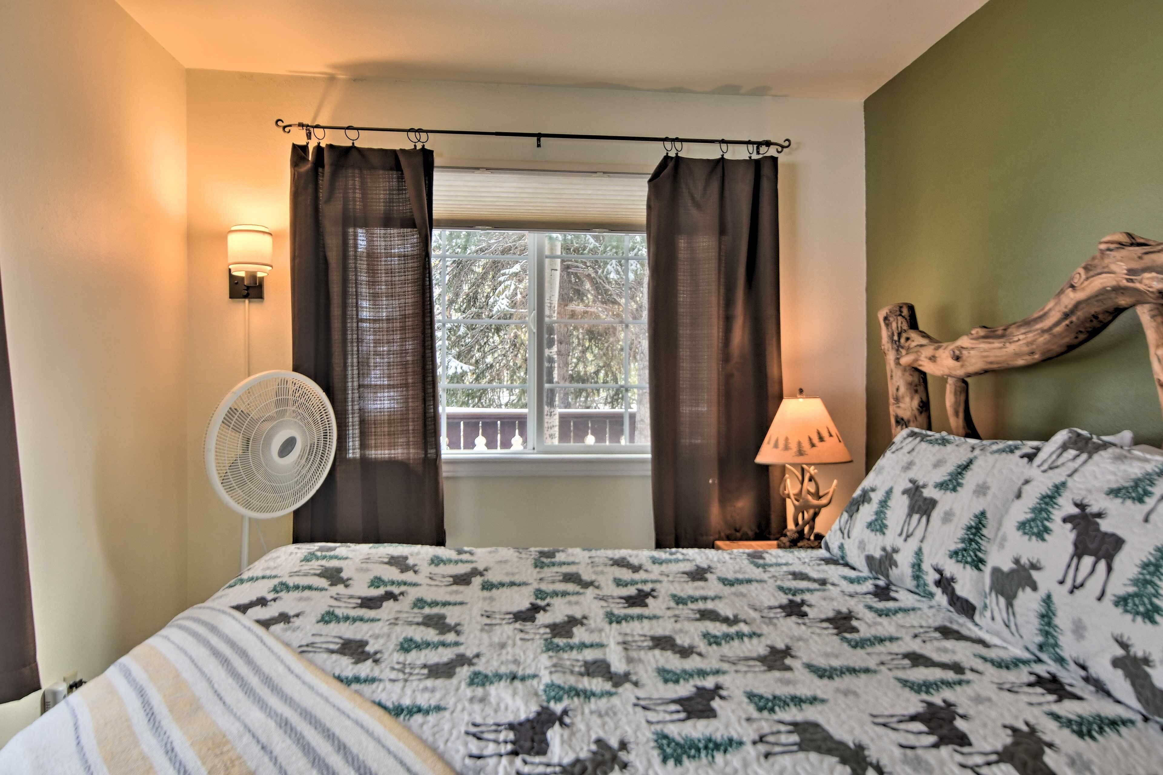 Un dormitorio con cama, ventilador de pie, ventana con cortinas, mesita de noche con lámpara y cabecera de madera.