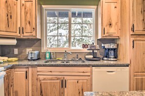 A kitchen with wooden cabinets, a window with a view of snow-covered trees, a toaster, a coffee maker, and a dishwasher. - Cozy Condo on Fall River - 1 Mile to Rmnp Gate! (Estes Park)