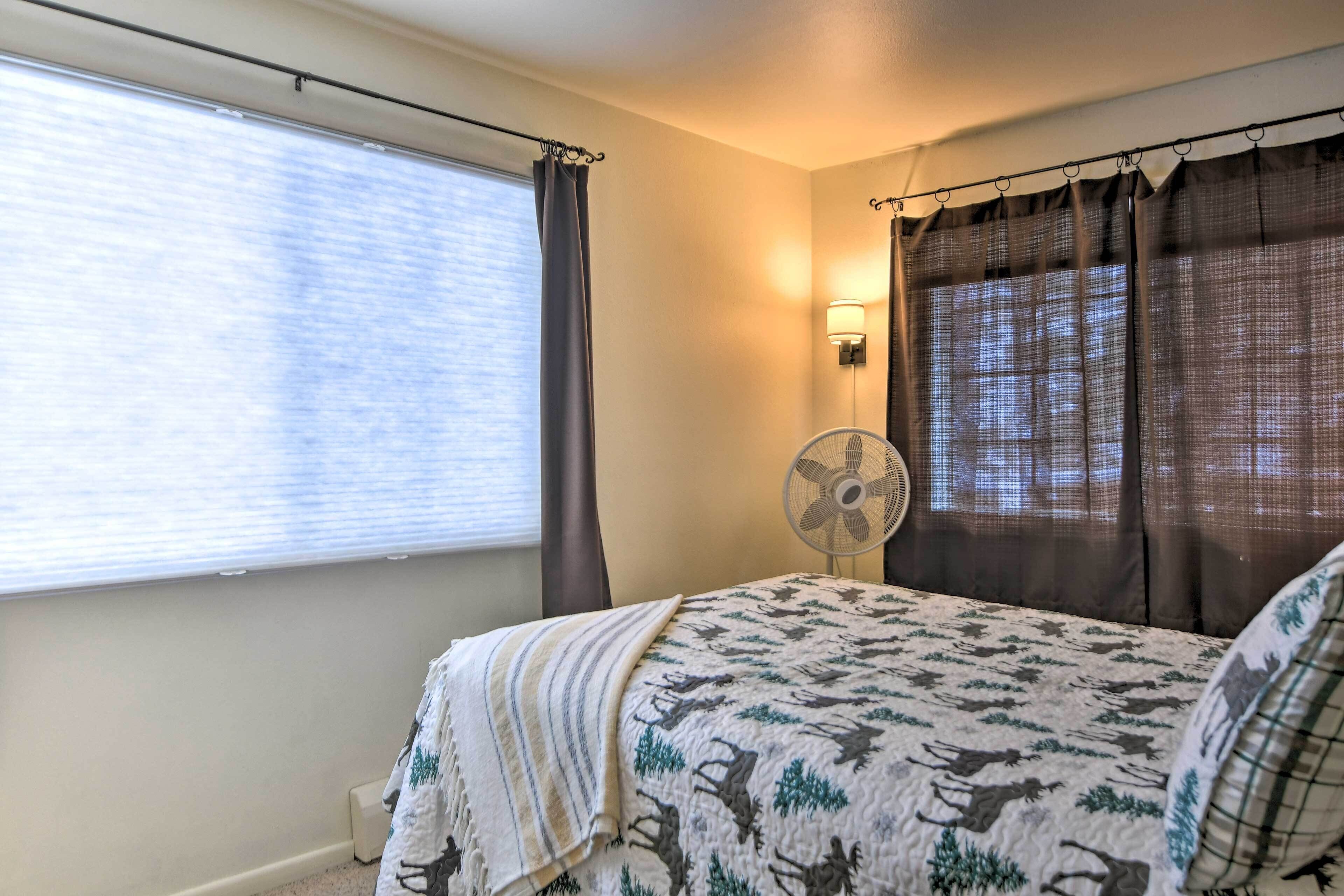 A bedroom with a bed, a window with curtains, a wall-mounted fan, and a wall-mounted light.