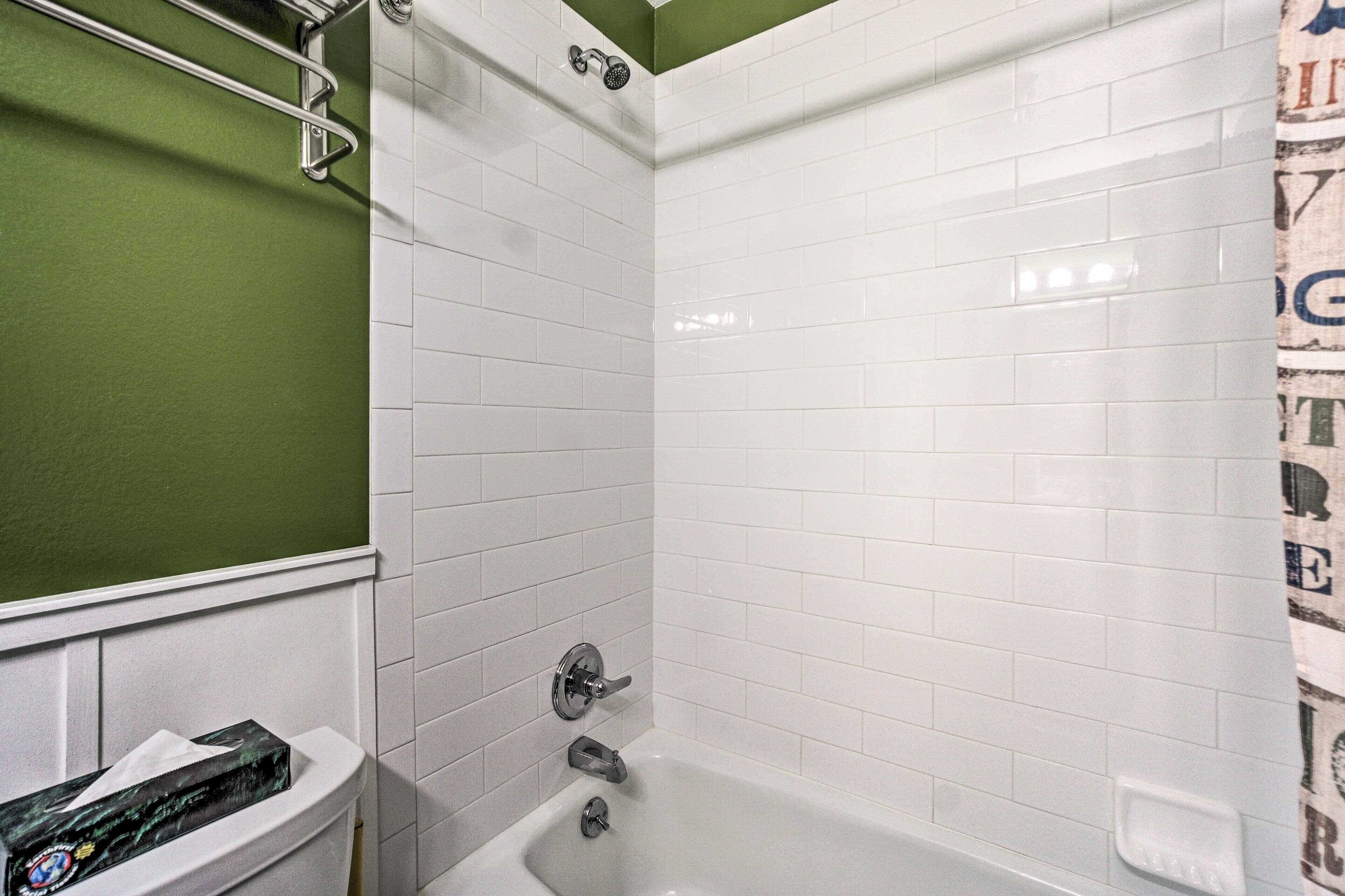 A bathroom with white tiled walls, a green accent wall, a white bathtub, a toilet with a box of tissues, and a shower area.