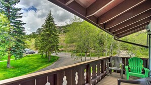 Une terrasse en bois dotée d’une rambarde, de chaises Adirondack vertes et offrant une vue sur un paysage verdoyant, avec des arbres et une route.