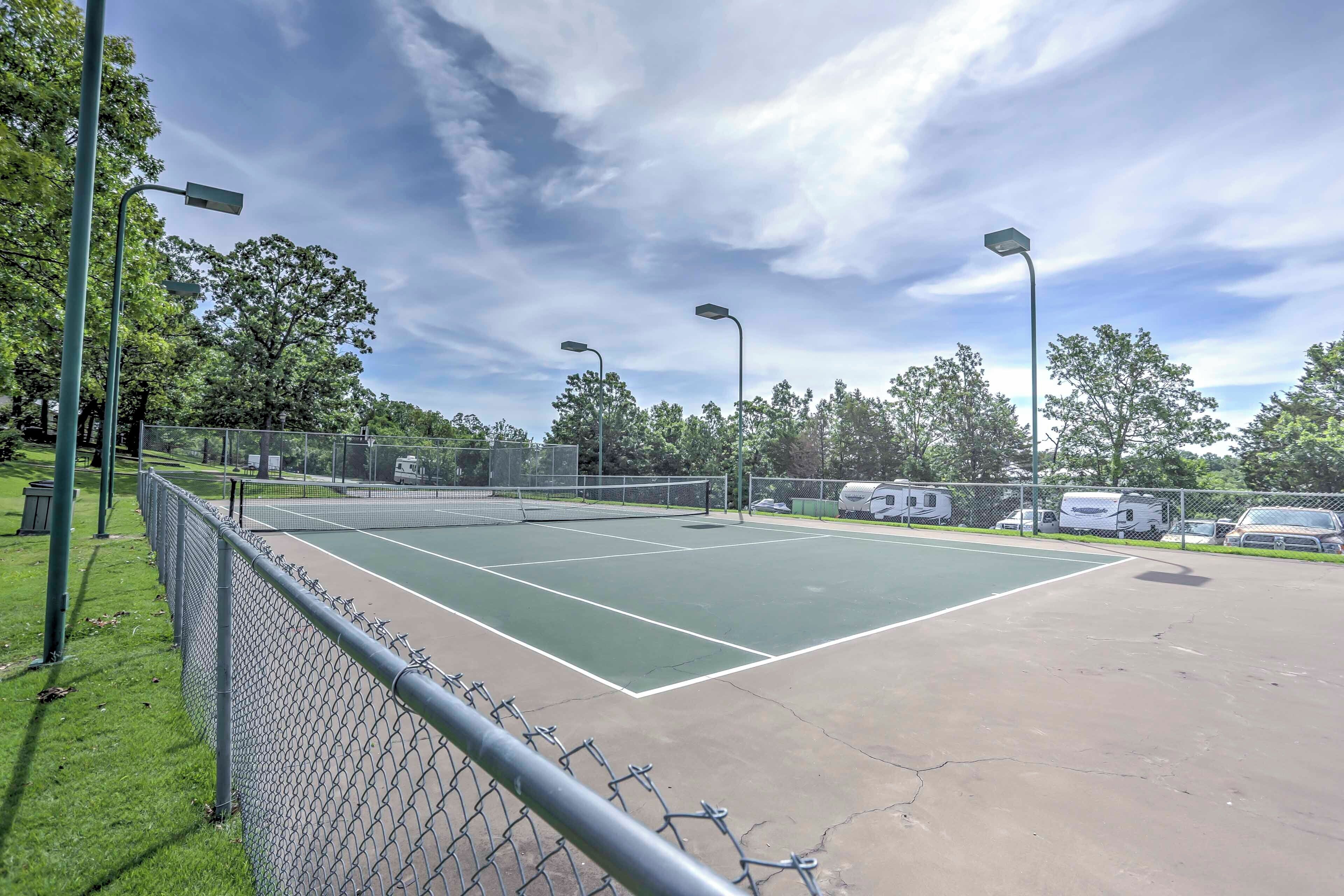 Una cancha de tenis al aire libre con una cerca de malla metálica, farolas y vehículos recreativos estacionados cerca.