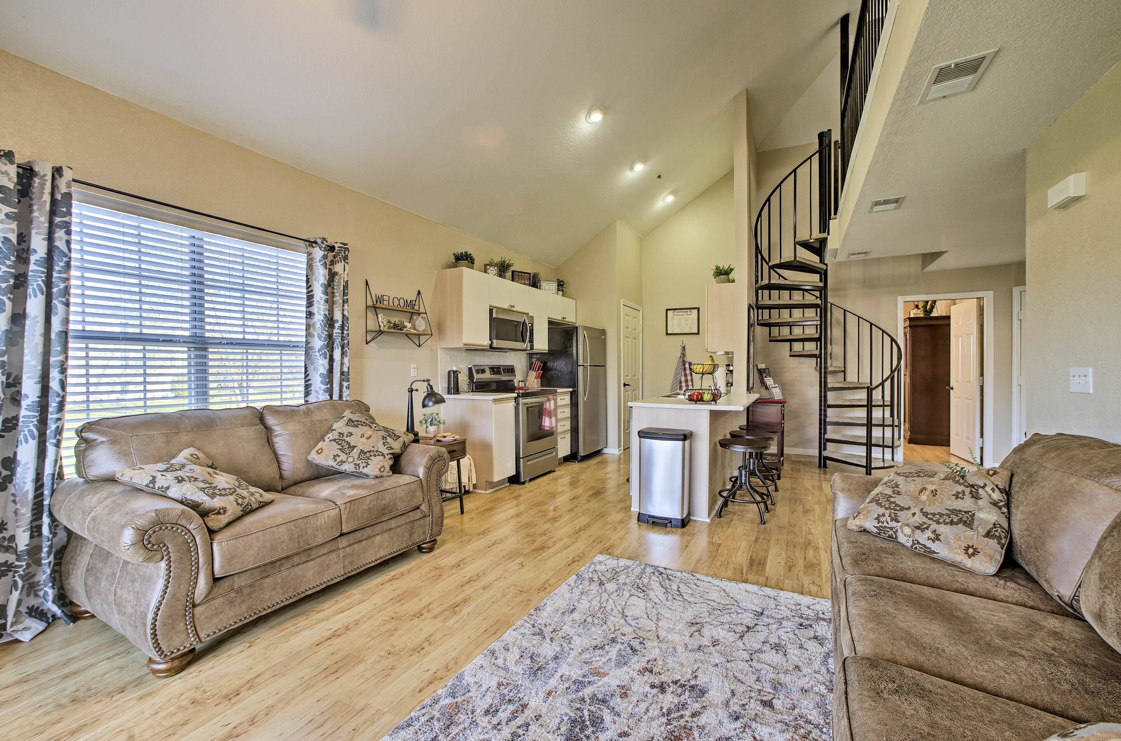 A spacious living room with a brown leather sofa, a coffee table, and a patterned rug. A kitchen with modern appliances and a staircase leading to an upper level is visible.