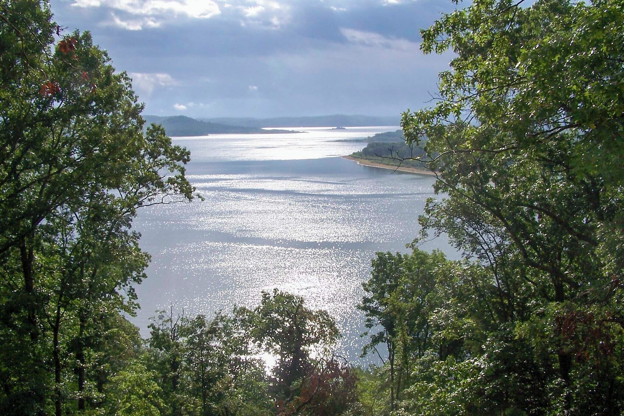A coastal landscape with a view of the sea and distant mountains, framed by dense green trees.