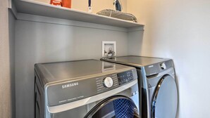 A Samsung washing machine and dryer in a laundry room with a shelf above containing a bottle, a knife, and a basket.