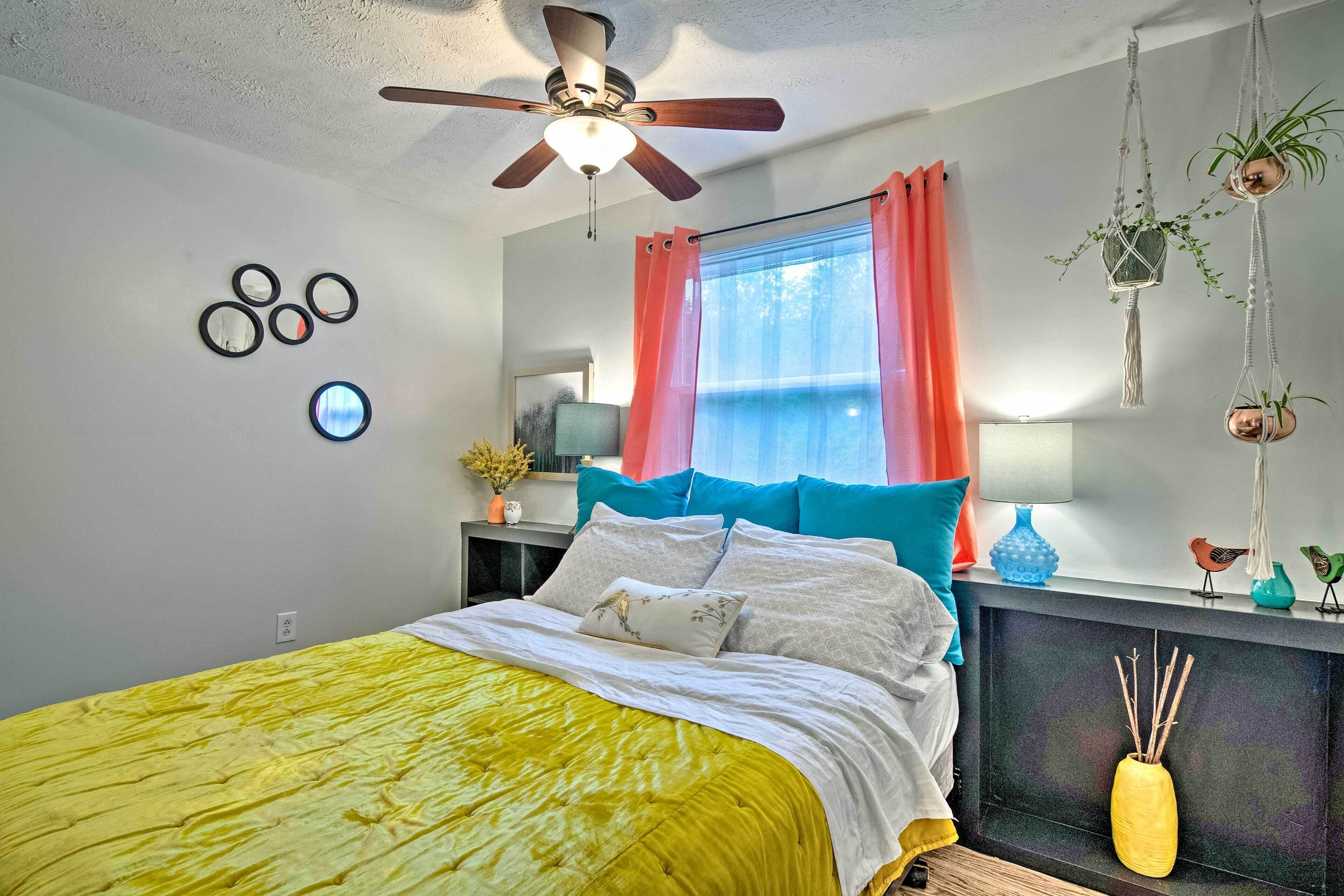 A bedroom with a bed, ceiling fan, window with curtains, bedside table, lamp, and wall decorations.