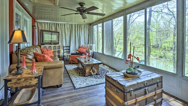 A cozy room with a wooden ceiling, a large window with a view of trees, a leather sofa, a wooden chest, and a table with a lamp. - Cozy Cabin With Sunroom & Cacapon River Access! (Capon Bridge)