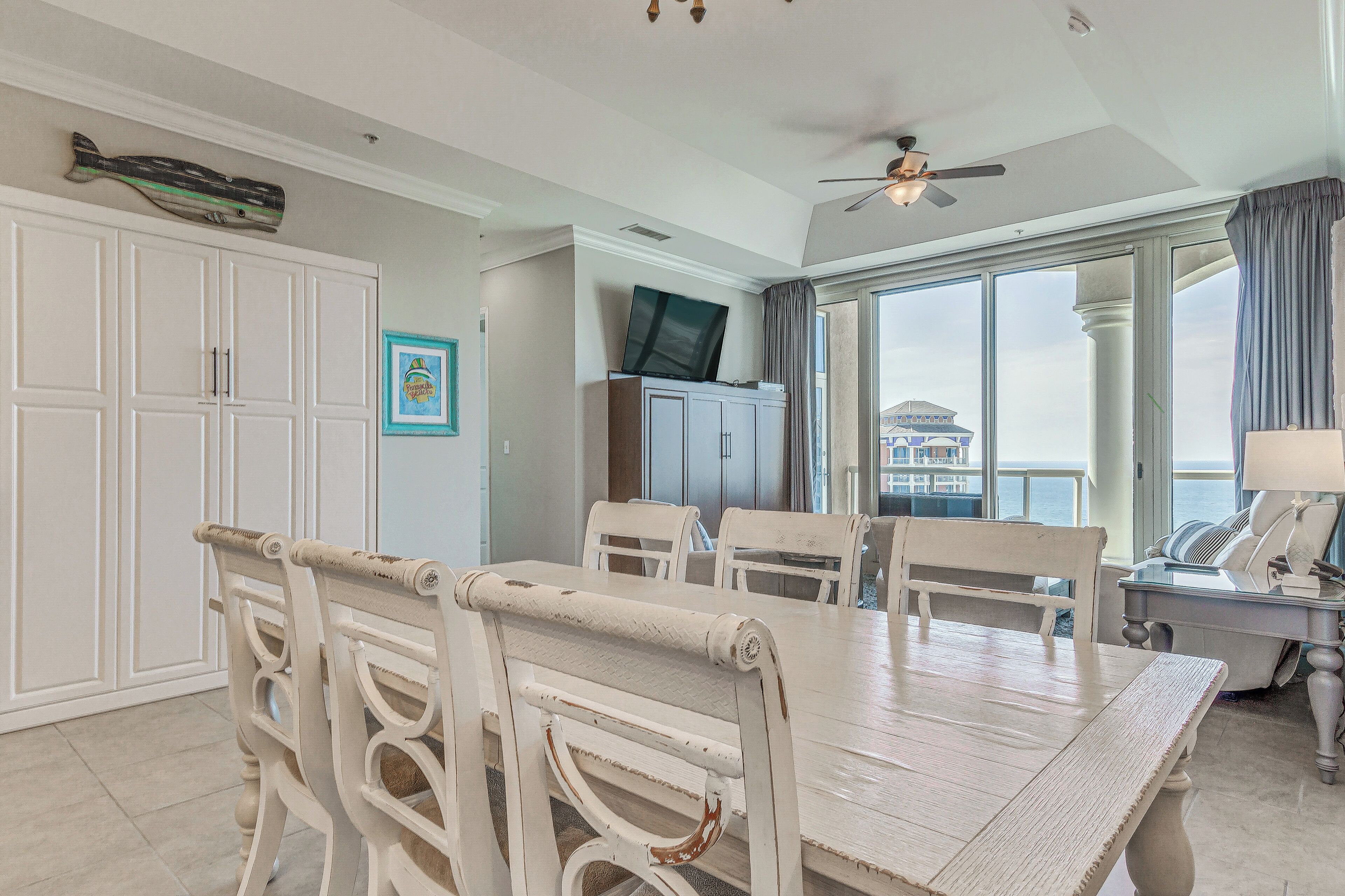A dining area with a white table and chairs, a ceiling fan, and a view of the ocean through large windows.