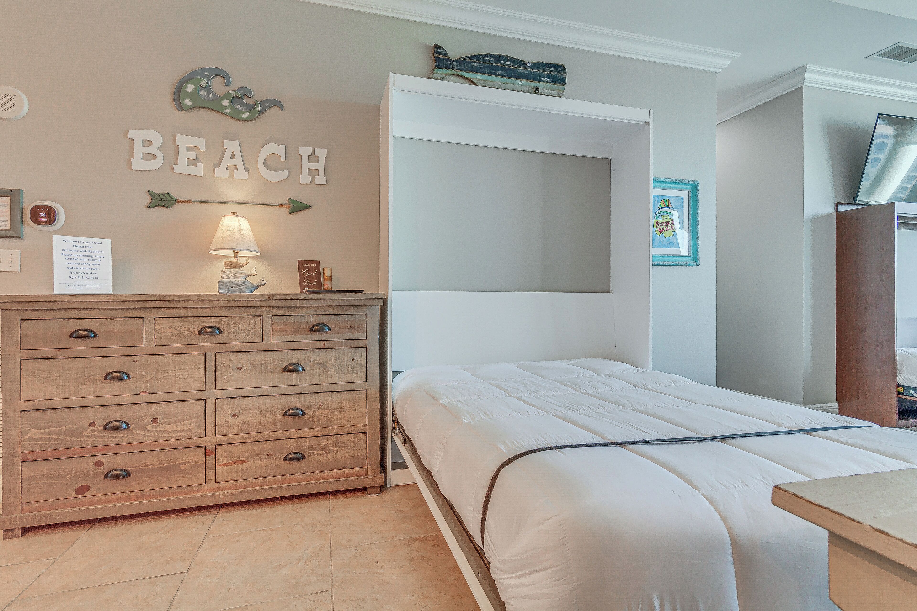A bedroom with a wooden dresser, a bed with white bedding, a wall-mounted shelf, and a sign that reads "BEACH".