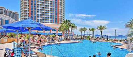A pool area with lounge chairs and umbrellas, surrounded by palm trees and a tall building in the background.