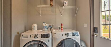 Un cuarto de lavandería con dos lavadoras de carga frontal, un estante con productos de limpieza y una ventana con vista al exterior.
