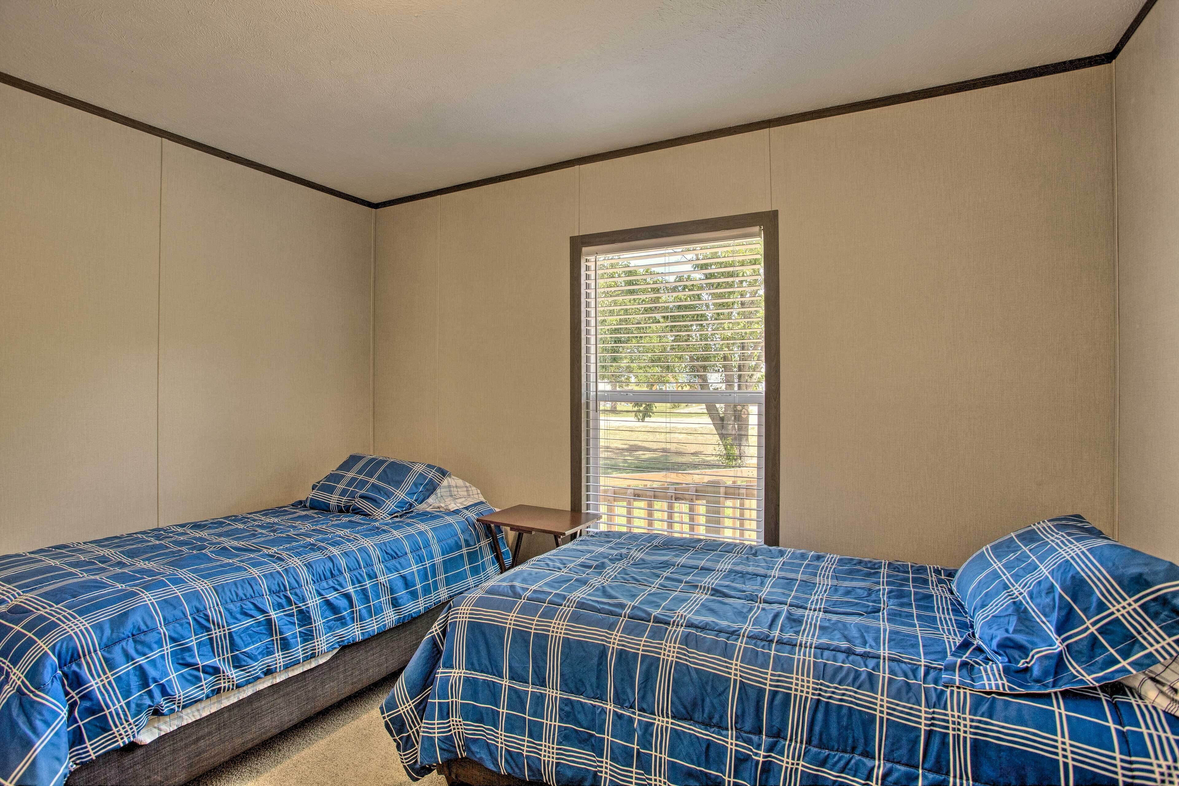 A room with two single beds, each with a blue checkered bedspread and matching pillows. There is a small wooden table between the beds. The room has beige walls and a window with blinds.