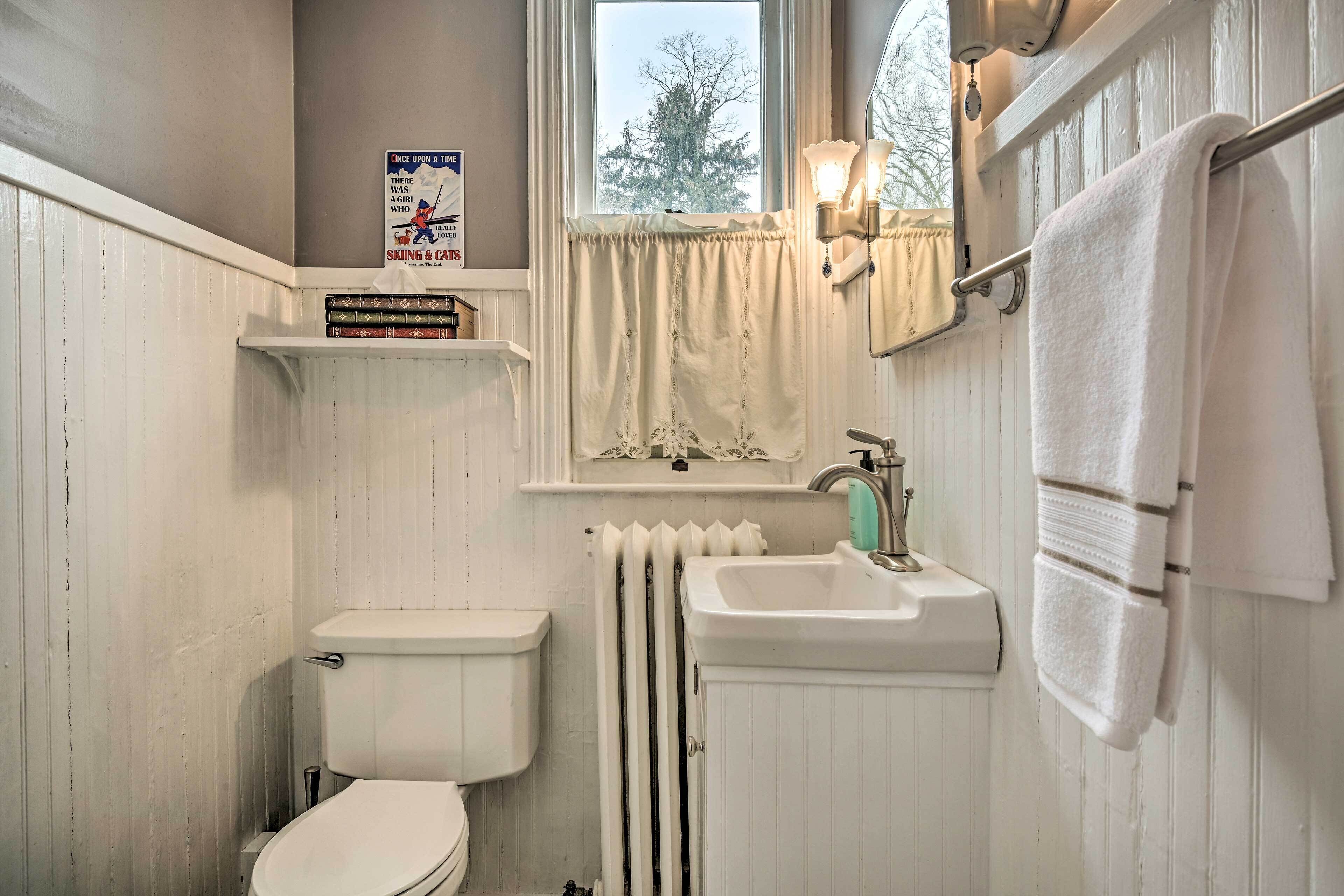 A compact bathroom with a toilet, sink, and radiator. There is a shelf with books and a sign above the toilet.