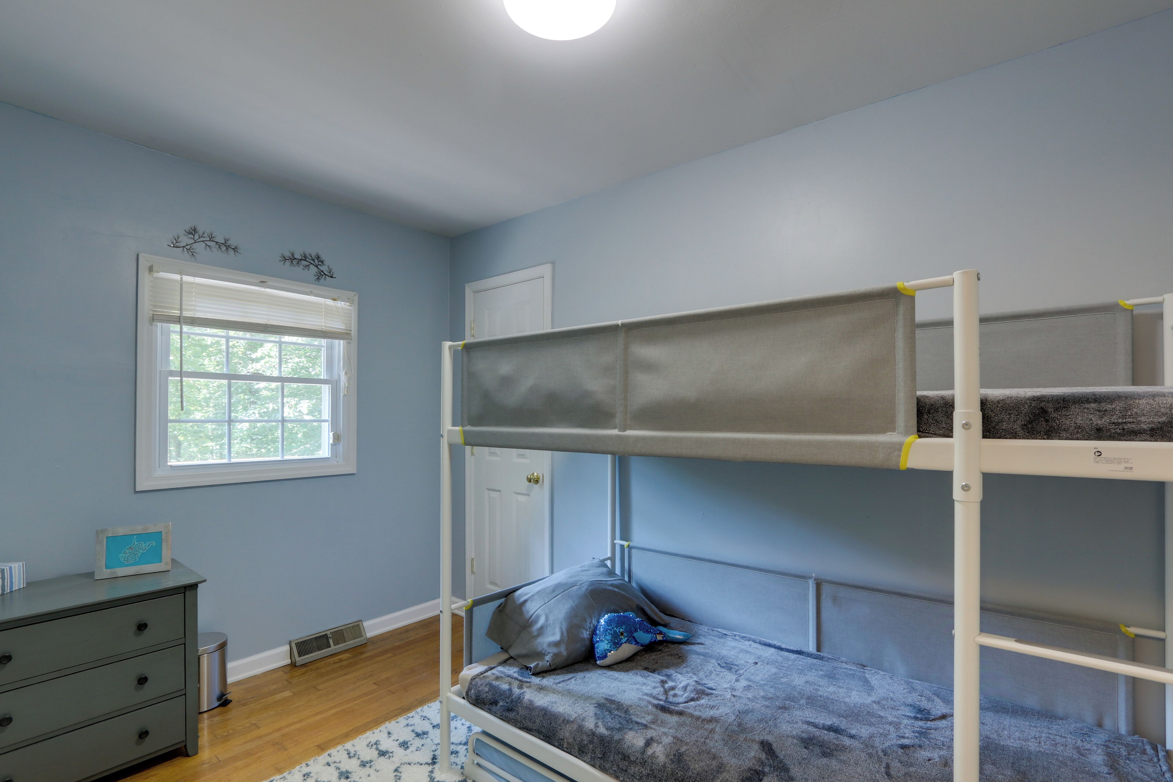 A bunk bed with a grey mattress and a pillow, a dresser, a window with blinds, and a door in a room with light blue walls.
